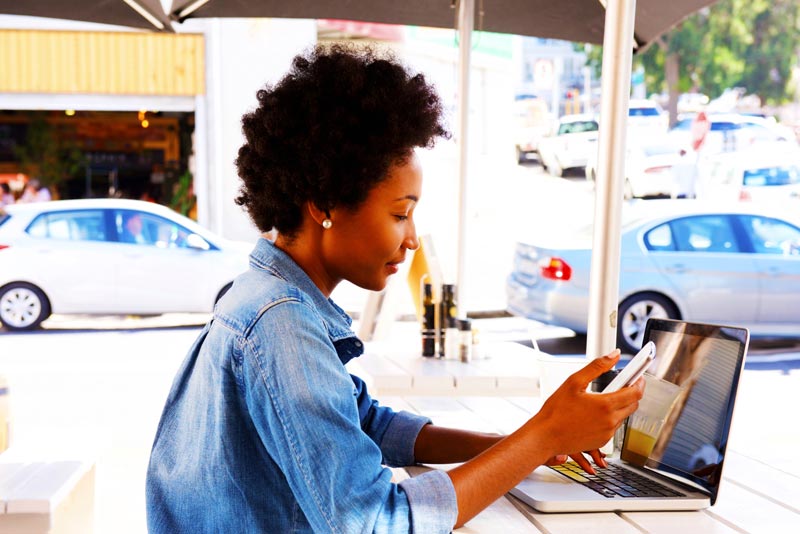 A woman city at a street side restaurant while working on her smart phone and laptop